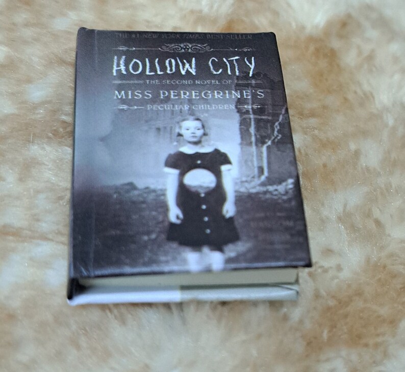 May include: A paperback book titled "Hollow City" by Ransom Riggs. The book is the second novel in the "Miss Peregrine's Peculiar Children" series. The cover features a black and white photograph of a young girl standing in front of a crumbling building.