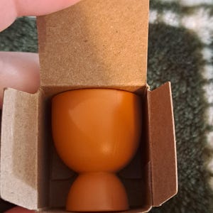 May include: A small, orange, ceramic egg cup. The cup is in a cardboard box.