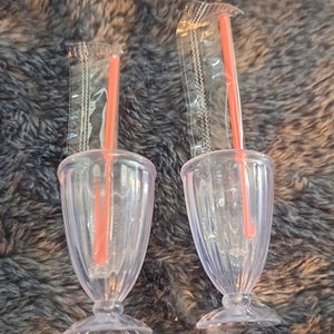 May include: Two clear plastic cups with red straws and clear plastic wrappers. The cups have a fluted design and a wide base.