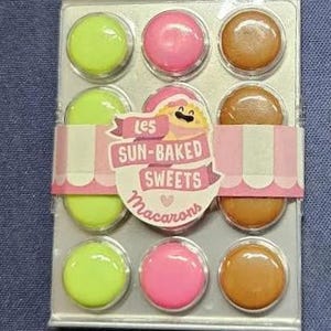 Pode incluir: Uma bandeja prateada com nove macarons coloridos. Os macarons estão dispostos em três filas de três. Os macarons são verdes, rosa e castanhos. A bandeja tem o texto "Les Sun-Baked Sweets Macarons" impresso nela.