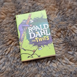以下が含まれることがあります： クエンティン・ブレイクが挿絵を描いた、ロアルド・ダールによる「The Twits」というタイトルの緑色のペーパーバック。表紙には、はしごに乗って緑色の背景を筆で塗っている男性と、長い腕と脚を持つ生き物の白黒のイラストが描かれている。