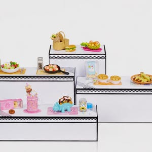 Puede incluir: Una colección de alimentos en miniatura, que incluye una ensalada, una hamburguesa y una tarta, dispuestos sobre plataformas blancas. Otros artículos incluyen un batido, un postre con forma de dinosaurio azul y un tarro de chispas. La escena se desarrolla sobre un fondo blanco.