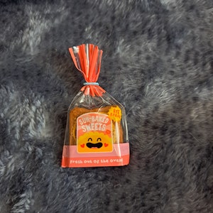 Puede incluir: Una bolsa de plástico transparente con un diseño rojo y blanco que contiene un pequeño juguete blando en forma de pan amarillo. El juguete tiene una cara sonriente y las palabras "Sun-Baked Sweets" y "Rye Bread" impresas en él. La bolsa también tiene el texto "Fresh out of the oven!" impreso en ella.