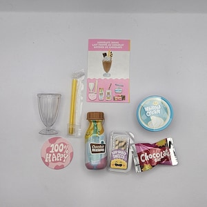 May include: A miniature set of chocolate milkshake ingredients, including a glass, a straw, a bottle of chocolate milkshake, a packet of sun-baked sweets, a container of whipped cream, and a chocolate bar. The set also includes a pink and white sticker that says "100% Happy".