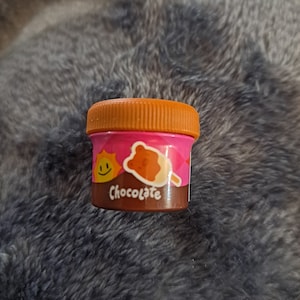 May include: A small, round, pink container with a brown lid. The container has a cartoon sun, a bear, and an ice cream cone on it. The text "Chocolate" is printed on the container.