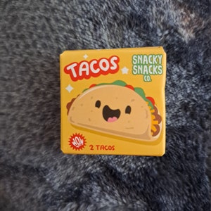Op de afbeelding: Een gele doos met het woord "TACOS" in rode letters en een cartoon taco-illustratie. De doos heeft ook de tekst "SNACKY SNACKS CO." en "NOW 2 TACOS". De achtergrond is een grijs, getextureerd oppervlak.