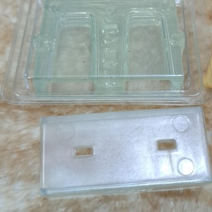 Może przedstawiać: Dwa przezroczyste plastikowe formy do tworzenia rękodzieła z żywicy. Formy są prostokątne i mają dwie wnęki. Formy leżą na jasnobrązowej powierzchni.