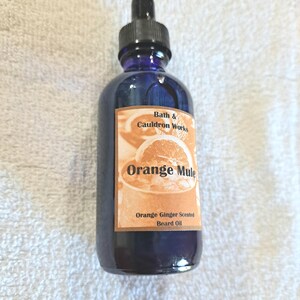 Puede incluir: Una botella de vidrio azul con un gotero, etiquetada "Bath & Cauldron Works Orange Mule Orange Ginger Scented Beard Oil".