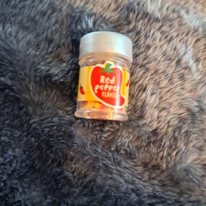 May include: A small clear jar with a white lid filled with red pepper flakes. The jar has a yellow label with a red pepper illustration and the text "Red Pepper Flakes".