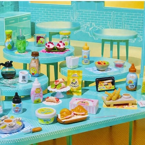 Puede incluir: Una exhibición de comida en miniatura con varios artículos como pasteles, bebidas y condimentos. La escena incluye pequeñas mesas y una encimera, todo en tonos azules y blancos. Los artículos incluyen una cafetera, una botella de crema y una bolsa de copos de tempura.