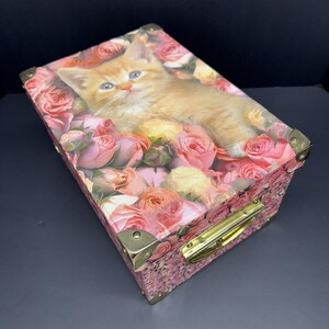 May include: A rectangular storage box featuring a printed image of an orange kitten resting among pink and peach roses. The box has metallic gold-toned corner reinforcements and a matching gold-toned metal handle on the side for carrying.