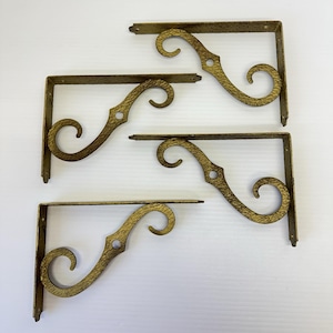 May include: A set of four L-shaped metal shelf brackets with a textured gold finish. Each bracket features a decorative scrollwork design with curved ends and a central circular hole for mounting, providing structural support.