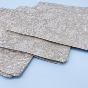 Puede incluir: Tres servilletas de tela beige dobladas con un estampado floral apiladas sobre una superficie azul claro. Las servilletas tienen una textura tejida y un brillo sutil, lo que sugiere un ambiente de comedor formal.