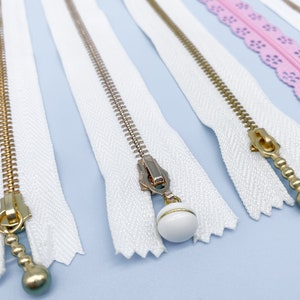 May include: Close-up of several zippers with white fabric tape and gold-colored metal teeth. Some zippers have decorative pulls, including a white bead and a gold-colored ball. A pink lace zipper is also visible.