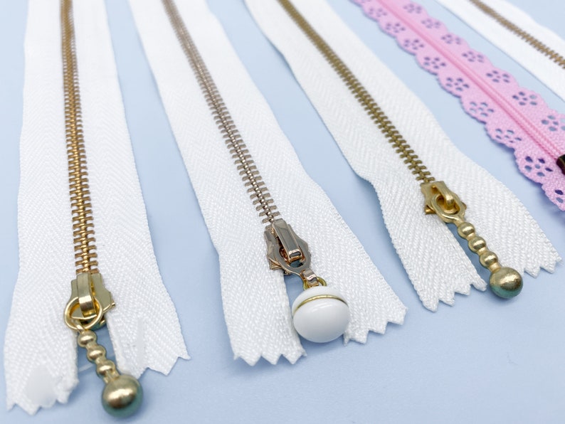 May include: Close-up of several zippers with white fabric tape and gold-colored metal teeth. Some zippers have decorative pulls, including a white bead and a gold-colored ball. A pink lace zipper is also visible.
