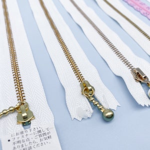 May include: Close-up of several white zippers with gold-colored metal teeth and pulls. Some pulls have decorative ball-shaped ends. The zippers are arranged on a light blue surface, showcasing their design and construction.
