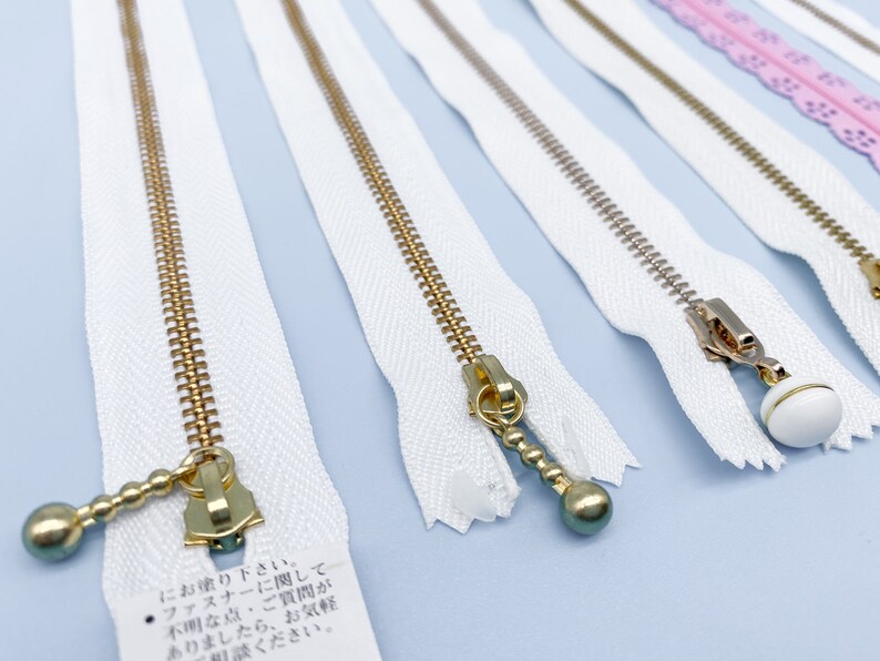 May include: Close-up of several white zippers with gold-colored metal teeth and pulls. Some pulls have decorative ball-shaped ends. The zippers are arranged on a light blue surface, showcasing their design and construction.