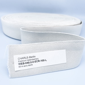 May include: A roll of white elastic band with a label that reads "CHARLE-Berlin Premium haberdashery WEB-040.0-0-ECR-100-L 50 m ecru leicht"