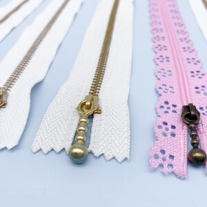 May include: Close-up of several zippers in various styles. White zippers with gold and silver metal teeth and pulls are displayed alongside a pink zipper with a decorative lace edge. The pulls have decorative ball and teardrop shapes.