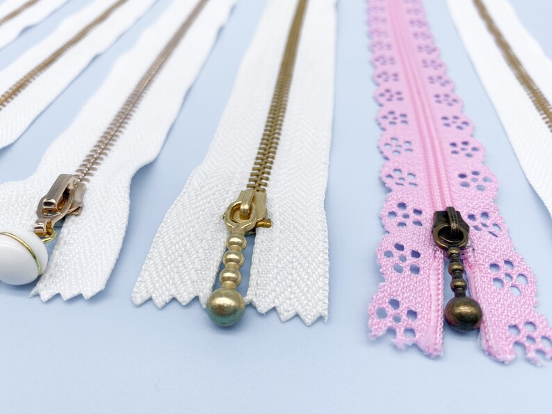 May include: Close-up of several zippers in various styles. White zippers with gold and silver metal teeth and pulls are displayed alongside a pink zipper with a decorative lace edge. The pulls have decorative ball and teardrop shapes.