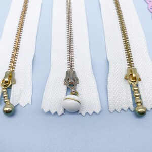 May include: Close-up of several white zippers with gold and silver metal teeth and pulls. One zipper has a white ball charm. A pink lace zipper is also visible. The zippers are on a light blue surface.