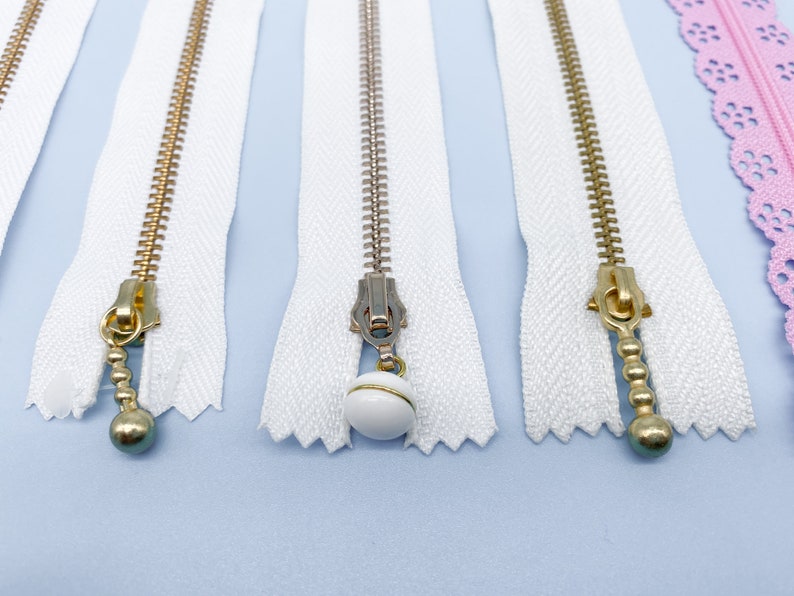 May include: Close-up of several white zippers with gold and silver metal teeth and pulls. One zipper has a white ball charm. A pink lace zipper is also visible. The zippers are on a light blue surface.
