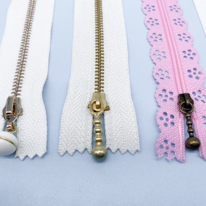 May include: Close-up of several zippers in various colors and styles. The zippers include white, pink, and gold-toned metal teeth. Some zippers have decorative pulls, including a white ball with gold trim and a pink lace edge.