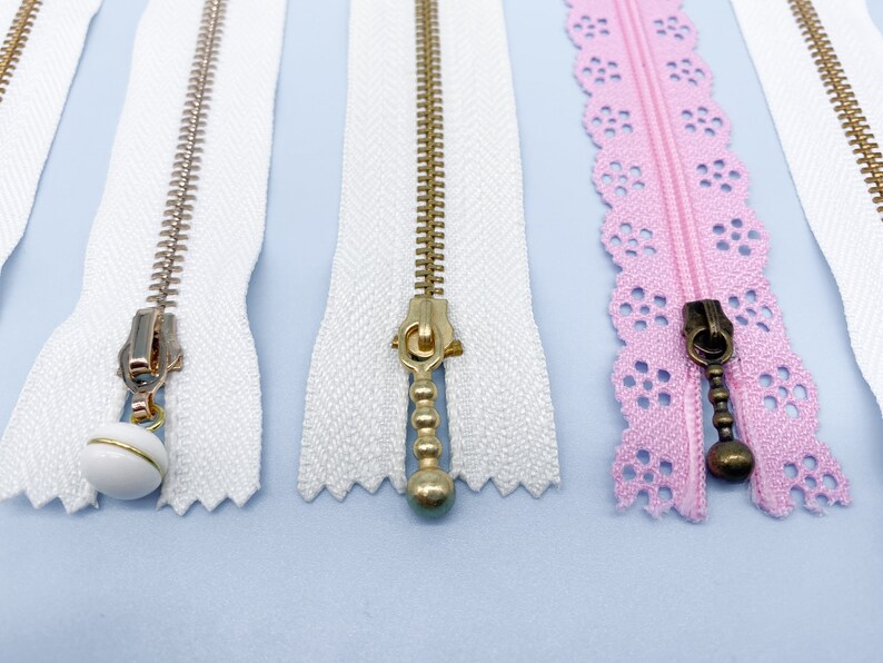 May include: Close-up of several zippers in various colors and styles. The zippers include white, pink, and gold-toned metal teeth. Some zippers have decorative pulls, including a white ball with gold trim and a pink lace edge.