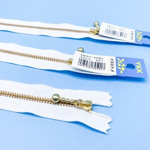 May include: Three white YKK zippers with gold-colored metal teeth and pulls. Each zipper has a small tag with Japanese text and a product code. The zippers are laid out on a light blue surface.
