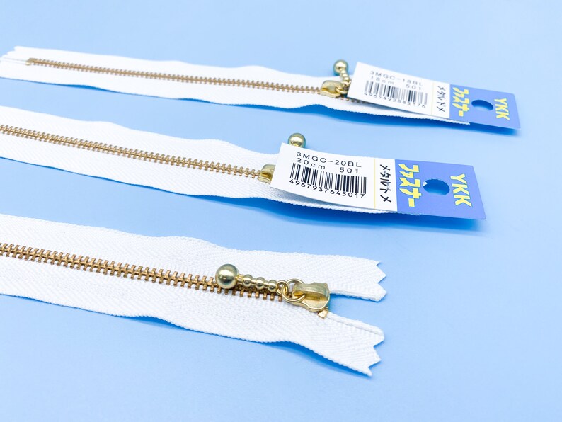 May include: Three white YKK zippers with gold-colored metal teeth and pulls. Each zipper has a small tag with Japanese text and a product code. The zippers are laid out on a light blue surface.