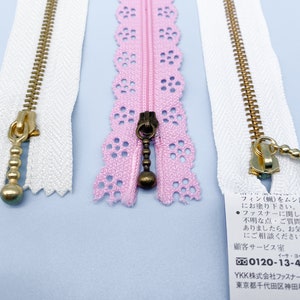 May include: Close-up of several zippers in various colors. Two white zippers with gold-colored pulls and teeth are visible, along with a pink lace zipper with a dark bronze pull. The zippers are on a light blue surface.