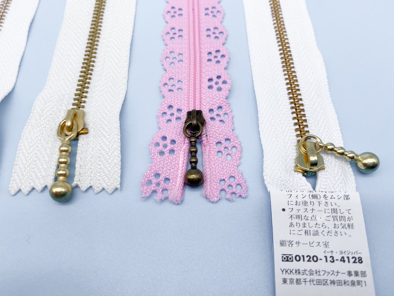 May include: Close-up of several zippers in various colors. Two white zippers with gold-colored pulls and teeth are visible, along with a pink lace zipper with a dark bronze pull. The zippers are on a light blue surface.