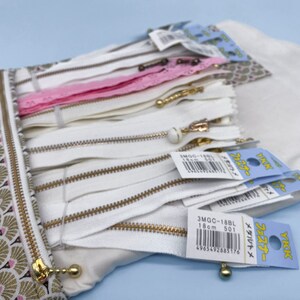 May include: A collection of white zippers with gold-colored pulls and tags, some with Japanese text. A pink lace zipper is also visible. The zippers are stored in a patterned pouch with a gold zipper closure. The pouch has a fan-like design in gold, pink, and brown.