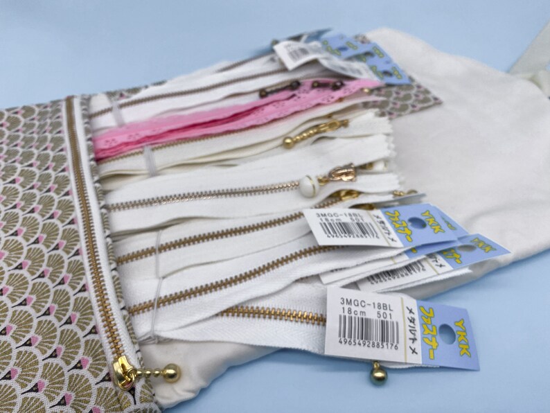 May include: A collection of white zippers with gold-colored pulls and tags, some with Japanese text. A pink lace zipper is also visible. The zippers are stored in a patterned pouch with a gold zipper closure. The pouch has a fan-like design in gold, pink, and brown.