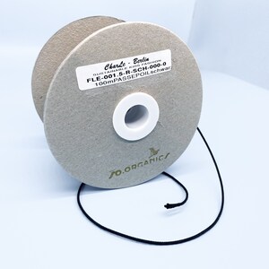 May include: A spool of black 100 meter passepoil trim, a type of fabric trim, on a cardboard spool with a white label that reads "CharLe-Berlia SUSTAINABLE KIDS FASHION FLE-001.5-R-SCH-000-0 100mPASSEPOIL.schwar" and a green logo that reads "FO.ORGANICS".