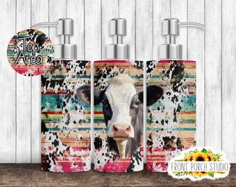 May include: Three silver soap dispensers with a cow print design. The design features a black and white cow head with a red, orange, yellow, blue, and green striped background. The dispensers are on a wooden surface.