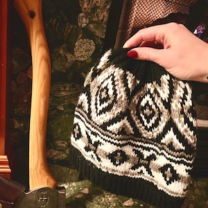 May include: A black and white knitted hat with a geometric pattern. The hat is folded and held by a hand with red nail polish.