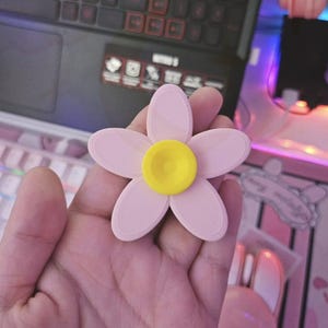 May include: A light pink, five-petal flower with a yellow centre. The flower is held in a hand, with a laptop and keyboard in the background. The flower appears to be 3D printed.