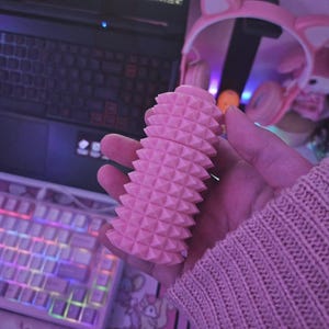 May include: A pink, textured, cylindrical object with a geometric pattern of raised triangles. The object is held in a hand, with a pink sweater sleeve visible. A laptop, keyboard, and pink cat ear headphones are in the background.