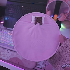 Könnte beinhalten: Ein rosa, rundes Plüschbett mit einem kleinen weißen Kissen und einer braunen Bärenfigur. Das Bett ist teilweise mit einer rosa Decke bedeckt. Das Bild hat ein niedliches, skurriles Thema.