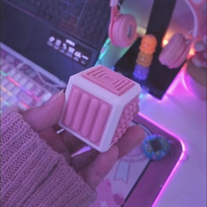 Pode incluir: Um cubo fidget rosa e branco é segurado em uma mão. O cubo tem várias texturas e designs em cada lado, incluindo uma série de cristas rosa, uma superfície texturizada e um padrão quadrado rebaixado. Outros itens no fundo incluem um laptop, fones de ouvido e um teclado.