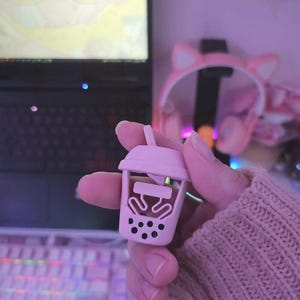 Puede incluir: Un dije en miniatura de taza de té boba de color rosa claro con una pajita. El dije tiene un diseño detallado, que incluye la taza, las perlas de boba y el té. El dije se sostiene en una mano, con un teclado y un portátil en el fondo.