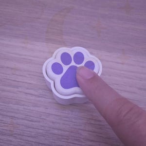 Op de afbeelding: Een witte pootafdrukstempel met een paars pootafdrukontwerp. Een vinger drukt op de paarse pootafdruk. De stempel ligt op een houten oppervlak.