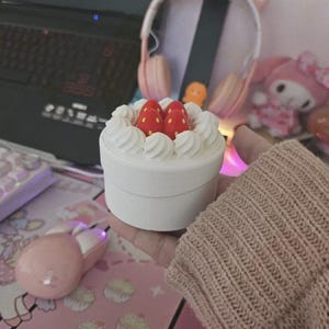 May include: A white, round container resembling a cake, topped with white frosting swirls and two red strawberry decorations. The container is held in a person's hand, with a pink mouse and keyboard visible in the background.
