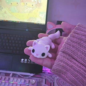 May include: A small, white cat figurine with black eyes and a pink nose is held in a hand. The cat is lying down with its tail up. A pink cat ear headband is visible in the background, along with a laptop and keyboard.