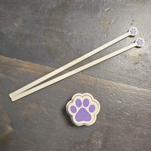 Può includere: Due bacchette bianche con dettagli a zampa viola e un sottobicchiere a forma di zampa abbinato. Le impronte delle zampe sono viola chiaro con un bordo bianco. Le bacchette sono sottili e dritte.
