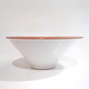 May include: A large, white ceramic bowl with a wide, terracotta-colored rim. The bowl has a smooth, glossy finish and a simple, modern design. The bowl is suitable for serving salads or as a decorative piece.