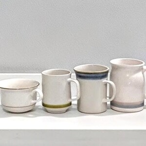 May include: Four ceramic mugs of varying sizes and designs. The mugs are off-white with subtle color accents, including green, blue, and a pinkish-gray. The mugs are arranged in a row, showcasing their different shapes and handles.