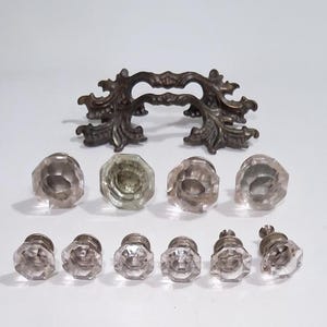 May include: An assortment of antique hardware, including a decorative bronze handle and several clear, faceted glass knobs. The knobs are arranged in two rows, with a variety of shapes and sizes. The hardware is set against a white background.