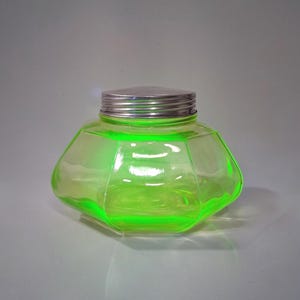 May include: A hexagonal, light green glass jar with a silver metal lid. The glass has a glowing, neon-like appearance. The jar is empty and sits on a white surface. The jar is likely vintage or antique.
